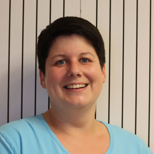 Eine lächelnde Person in blauer Kleidung vor einem gestreiften Hintergrund.