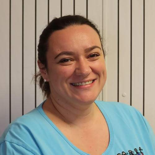 Eine lächelnde Person mit dunklem Haar, das zu einem Knoten gebunden ist, trägt ein hellblaues Oberteil vor einem Hintergrund mit vertikalen Streifen.