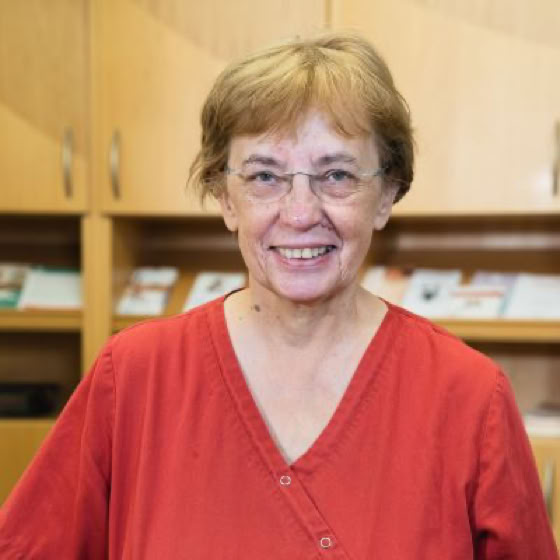 Eine lächelnde Person mit Brille und rotem Hemd steht vor einem Bücherregal.