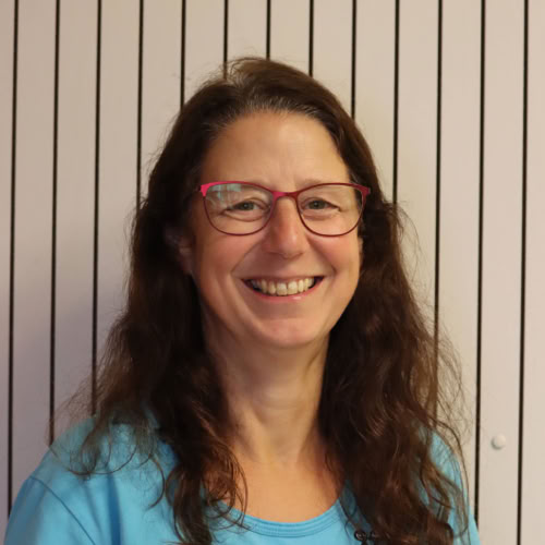 Eine Person mit Brille und blauem Oberteil steht vor einem Hintergrund mit vertikalen Streifen.