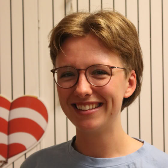 Eine Person mit einem weißen Kopftuch und einem roten Hemd steht vor einem bunten Hintergrund.