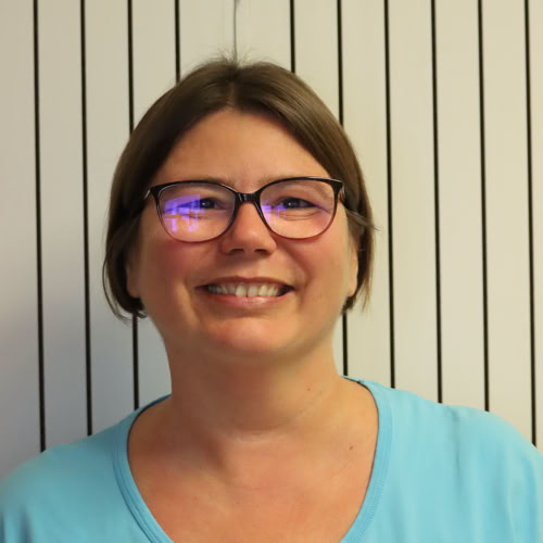 Eine lächelnde Person mit Brille in blauer medizinischer Kleidung steht vor einem Holzregal mit Broschüren.