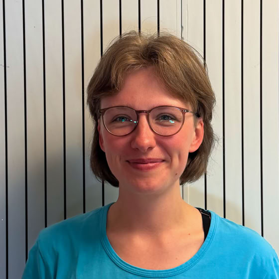 Eine Person mit einem weißen Kopftuch und einem roten Hemd steht vor einem bunten Hintergrund.