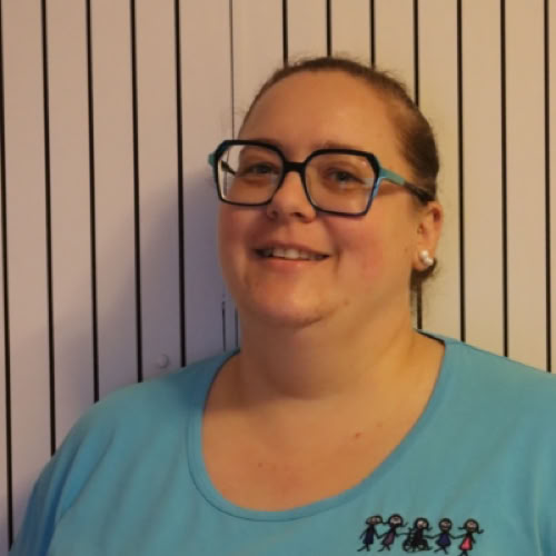 Eine Person mit Brille und blauem Oberteil steht vor einem Hintergrund mit vertikalen Streifen.